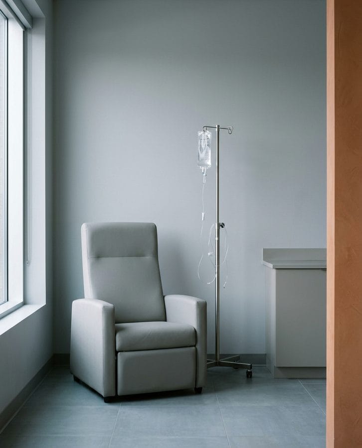 A plain upholstered armchair beside a stainless stand carrying one clear unlabelled fluid bag.