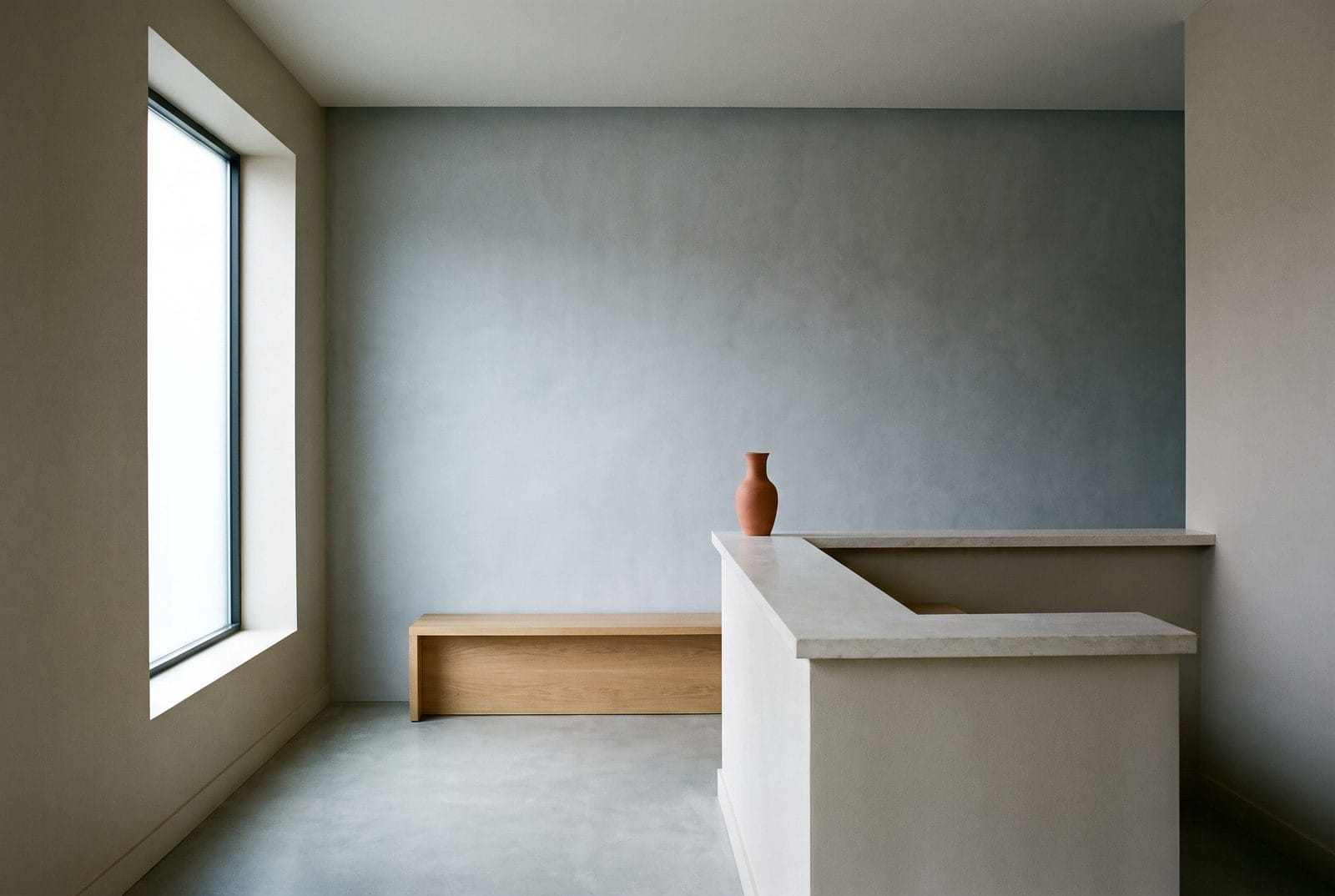 A reception counter in pale stone with nothing on it, a timber bench behind, lit from a tall window at one side.