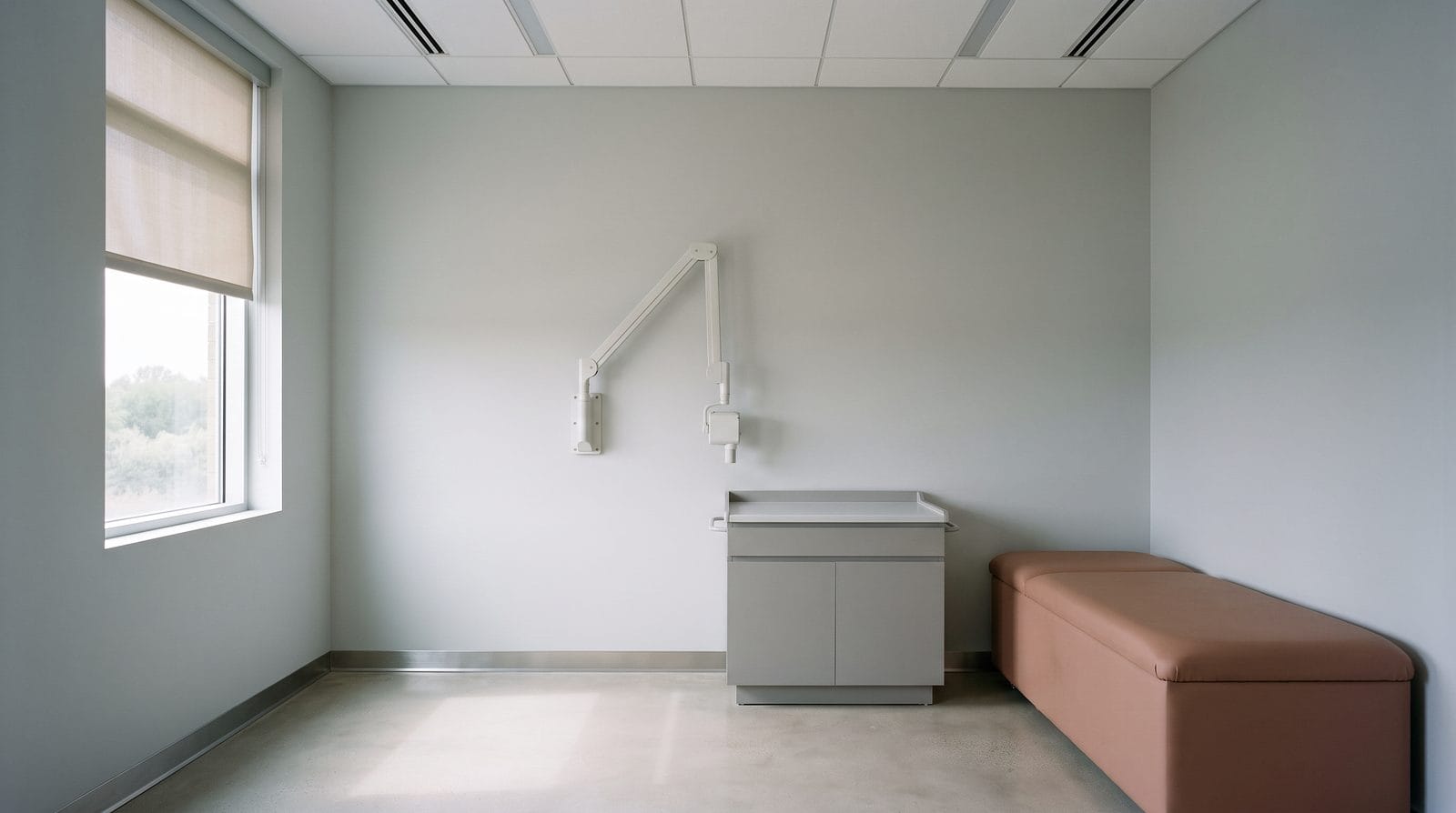 An empty clinical room with an articulated equipment arm folded against the wall and a half-drawn blind.