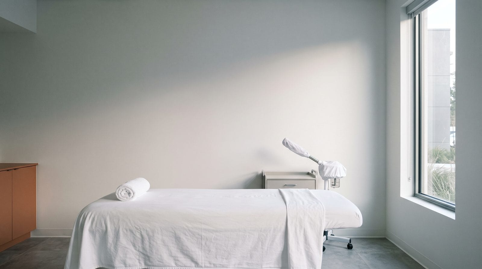 An empty facial room with a flat bed in fresh linen, a rolled towel at the head and a covered steam arm folded beside it.