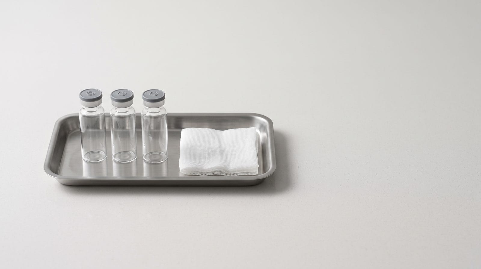 Three plain unlabeled glass vials standing in a row on a shallow stainless tray beside folded gauze.