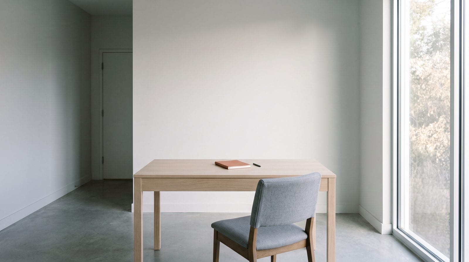 A plain desk against a pale wall with one chair pulled up to it and a closed notebook on top.