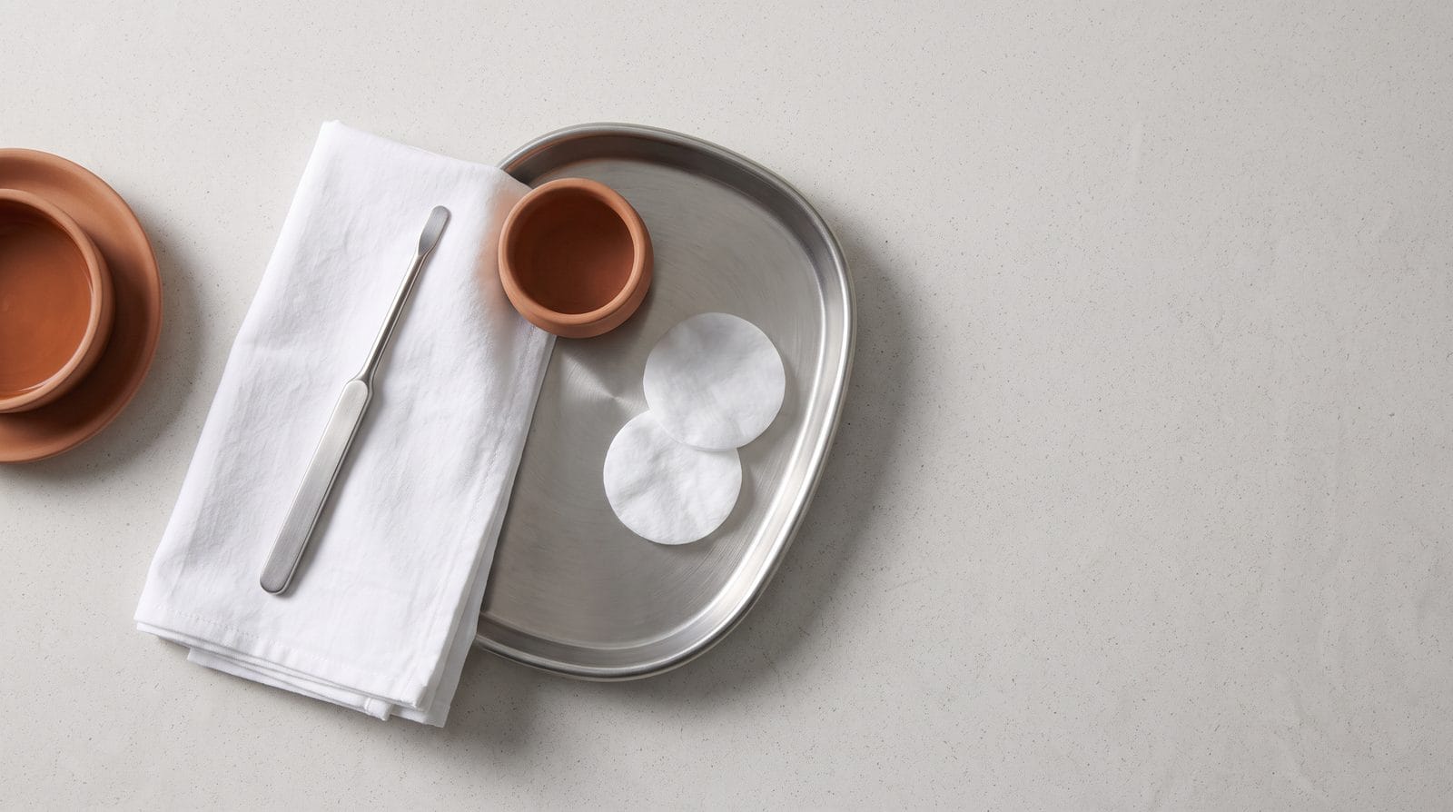 A plain stainless handle instrument lying on a folded white cloth beside a shallow tray and two cotton rounds.